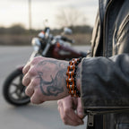 Person wearing a leather jacket and chain bracelet with a tattoo on their arm, standing next to a motorcycle.